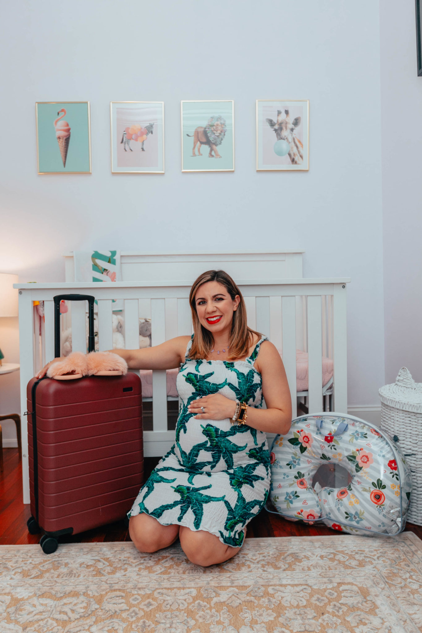 My Hospital Bag Essentials: What I Actually Used by popular Chicago lifestyle blog, Glass of Glam: image of a woman kneeling on the ground in her baby nursery next to a Boppy pillow and a rolling suitcase.
