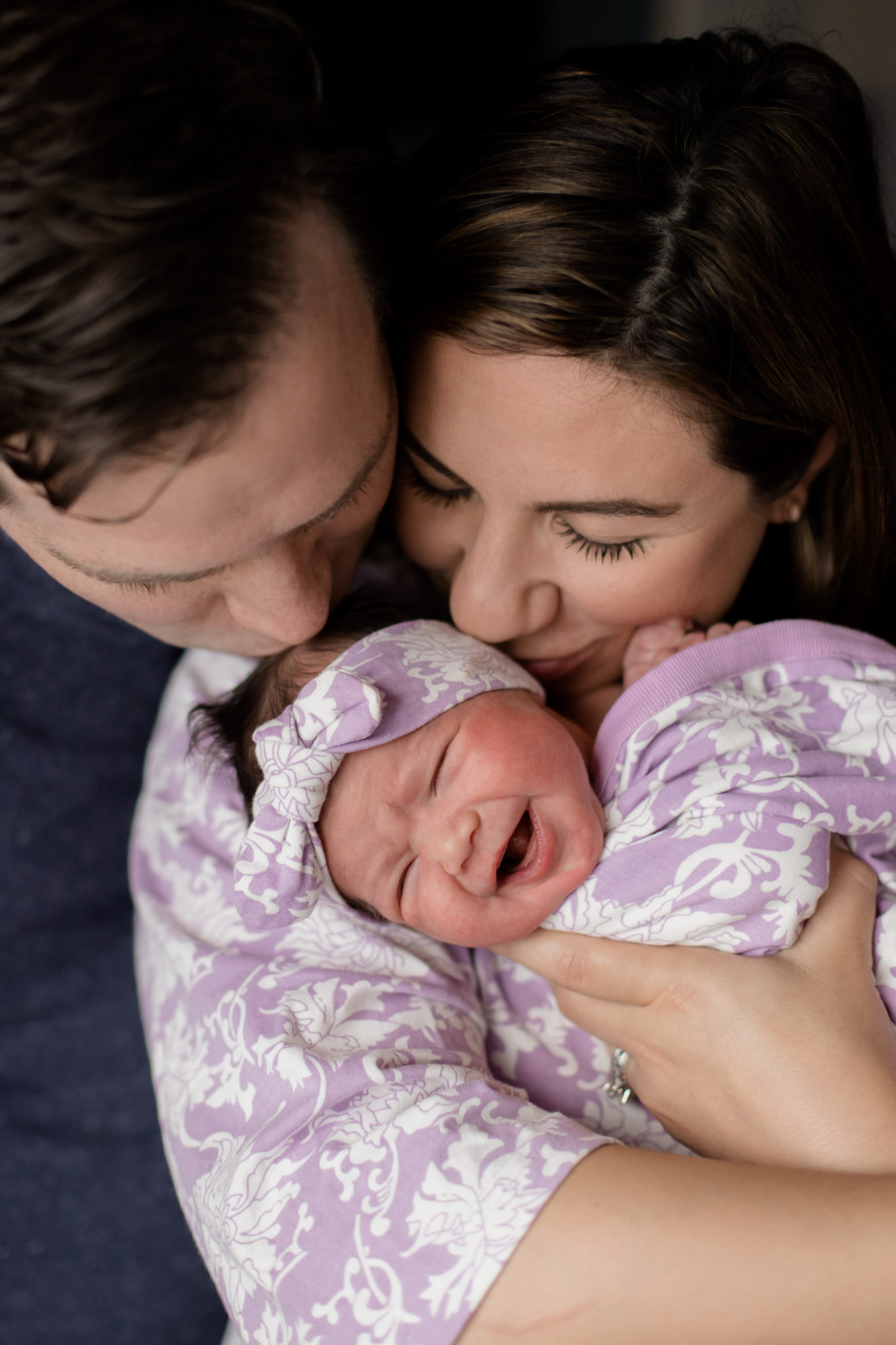 Baby Girl Birth Story: Welcome Sidney Eileen! by popular life and style blog, Glass of Glam: image of a man and woman sitting closely together and holding a newborn baby girl wrapped in purple and white floral print blanket.