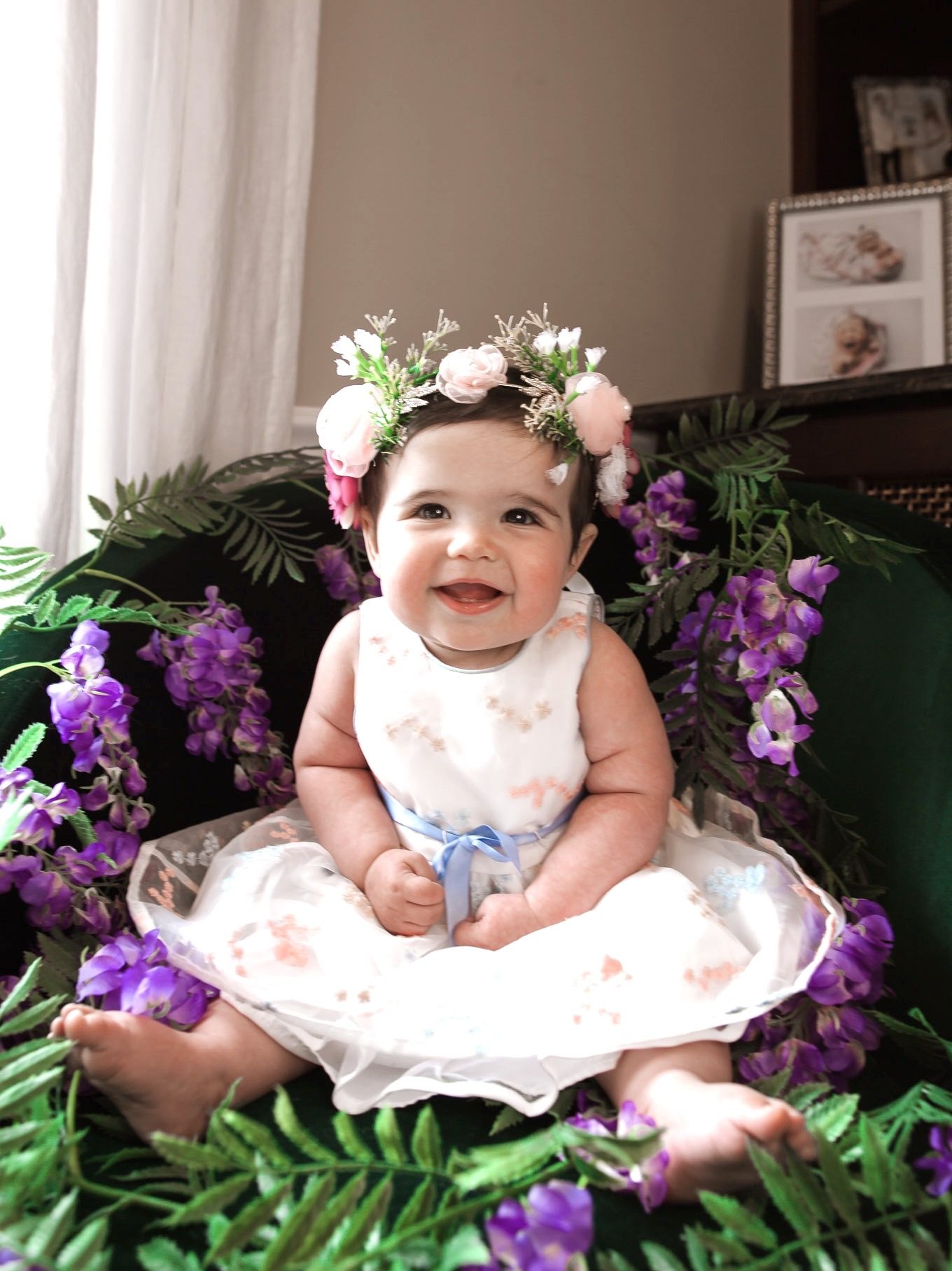 First Birthday by popular Chicago motherhood blog, Glass of Glam: image of a 9 month old baby girl wearing a white floral dress, flower crown and sitting in a wisteria garland.