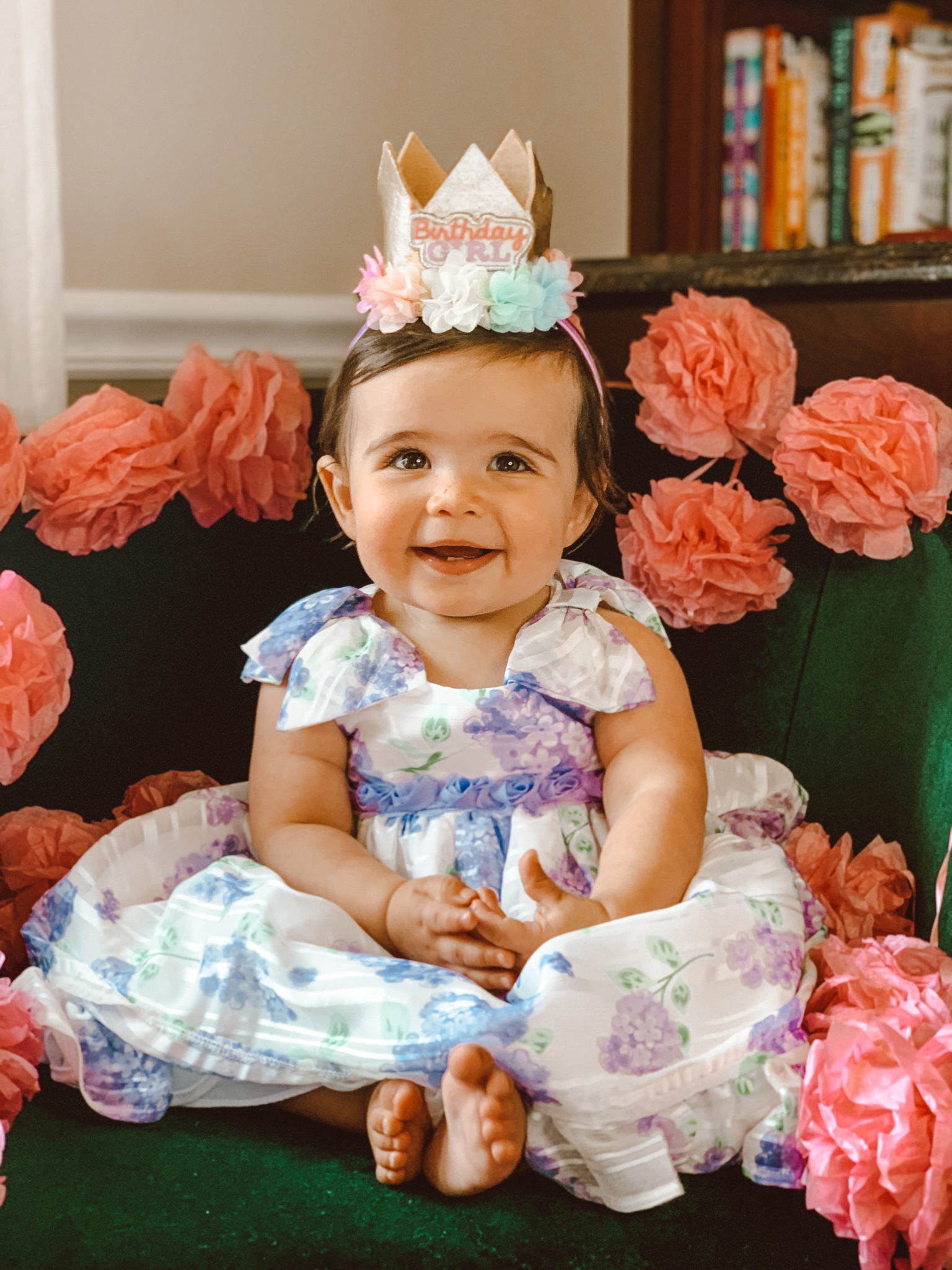 First Birthday by popular Chicago motherhood blog, Glass of Glam: image of a baby sitting on a green velvet chair surrounded by a tissue paper pom pom garland and wearing a white, blue and purple floral dress and gold 'Birthday Girl' crown. 