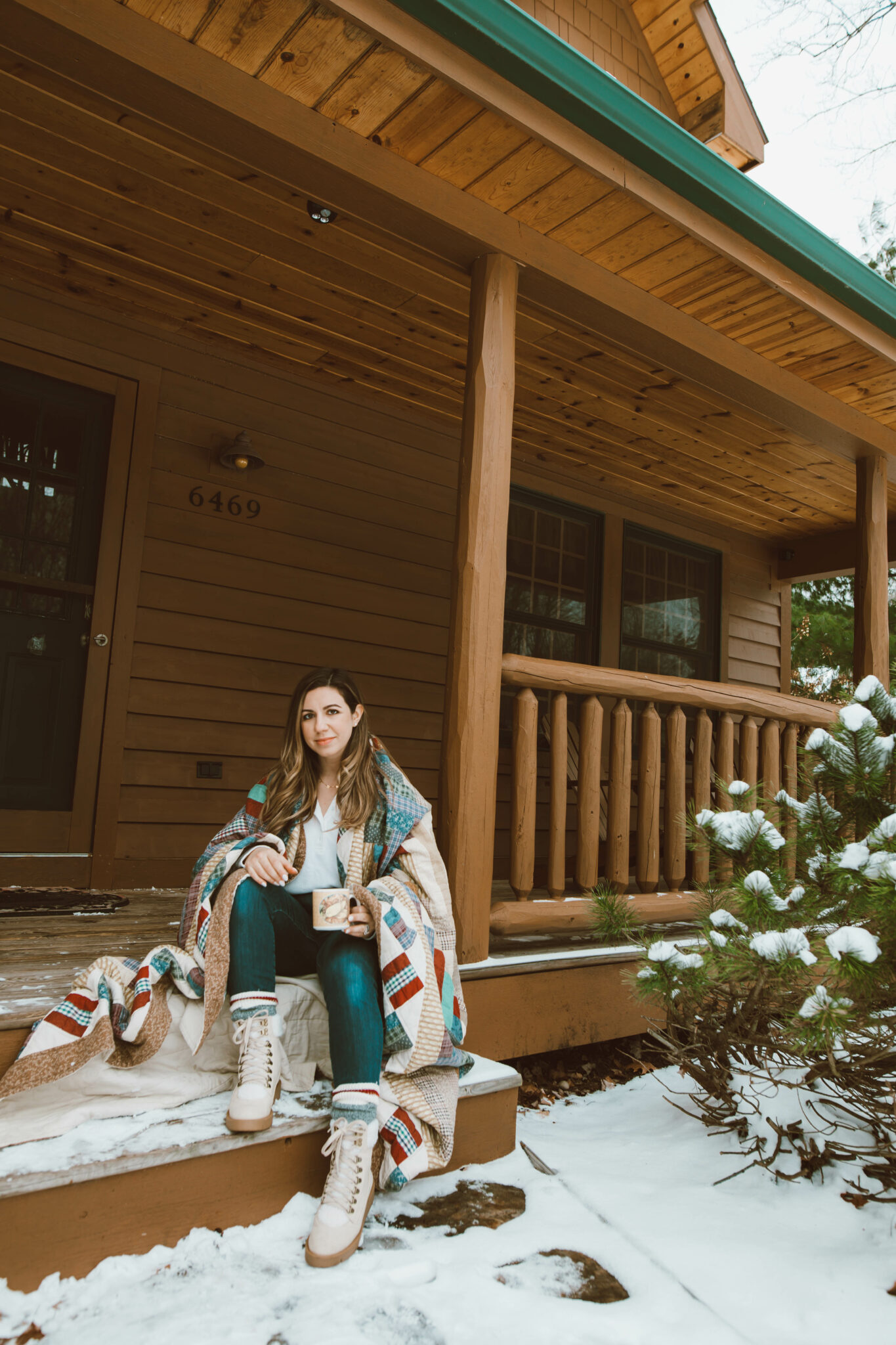 Fashion Basics by popular Chicago fashion blog, Glass of Glam: image of a woman sitting on outside of her cabin on the steps of her front porched wrapped up in a blanket and wearing a white henley t-shirt, jeans, JustFab hiking boots, and cabin socks. 