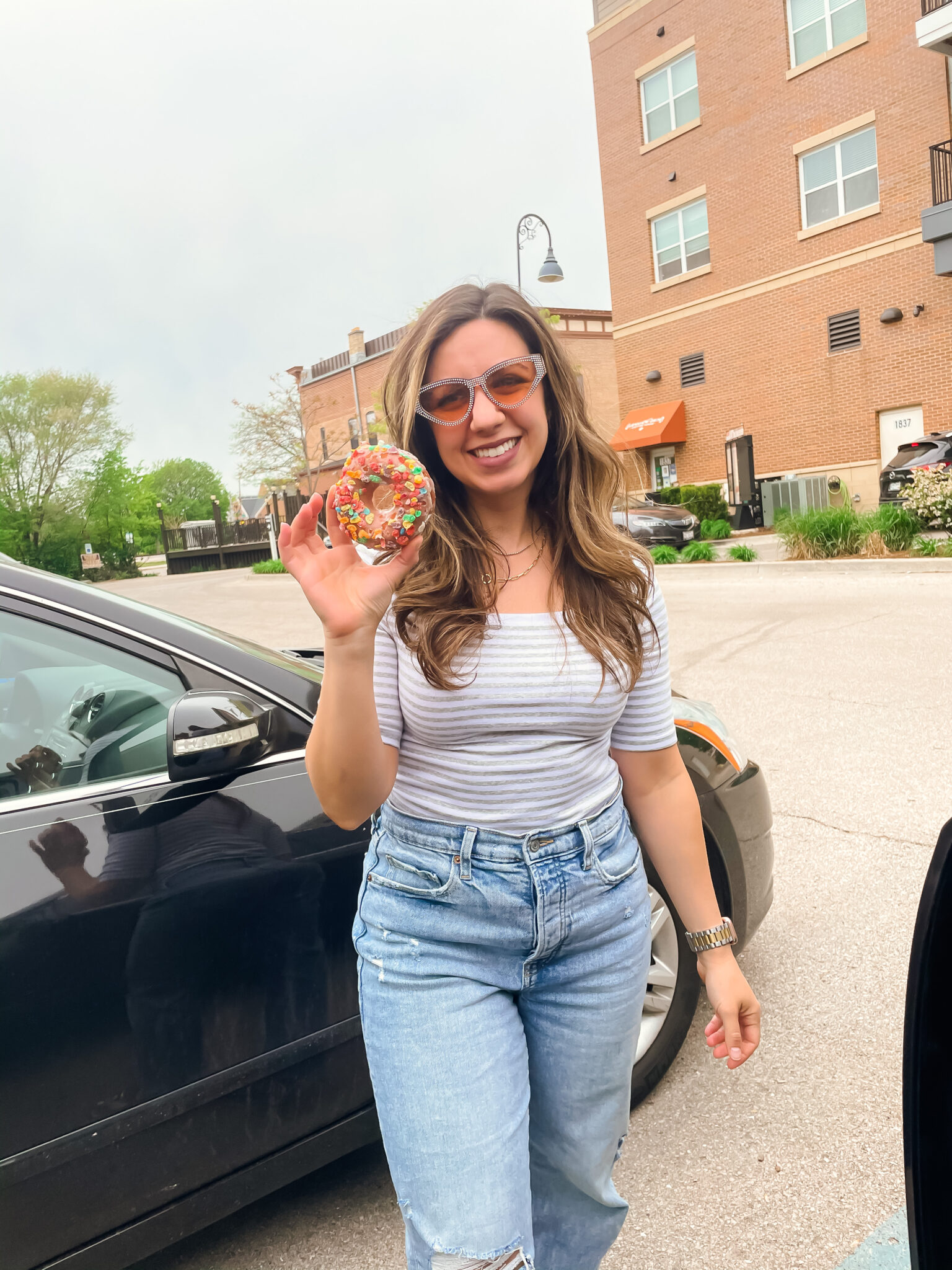 Amazon Favorites by popular Chicago life and style blog, Glass of Glam: image of a woman standing in a parking lot next to a black sedan and holding a donut with fruity pebbles on it and wearing a grey and white strip shirt with a pair of light wash jeans. 