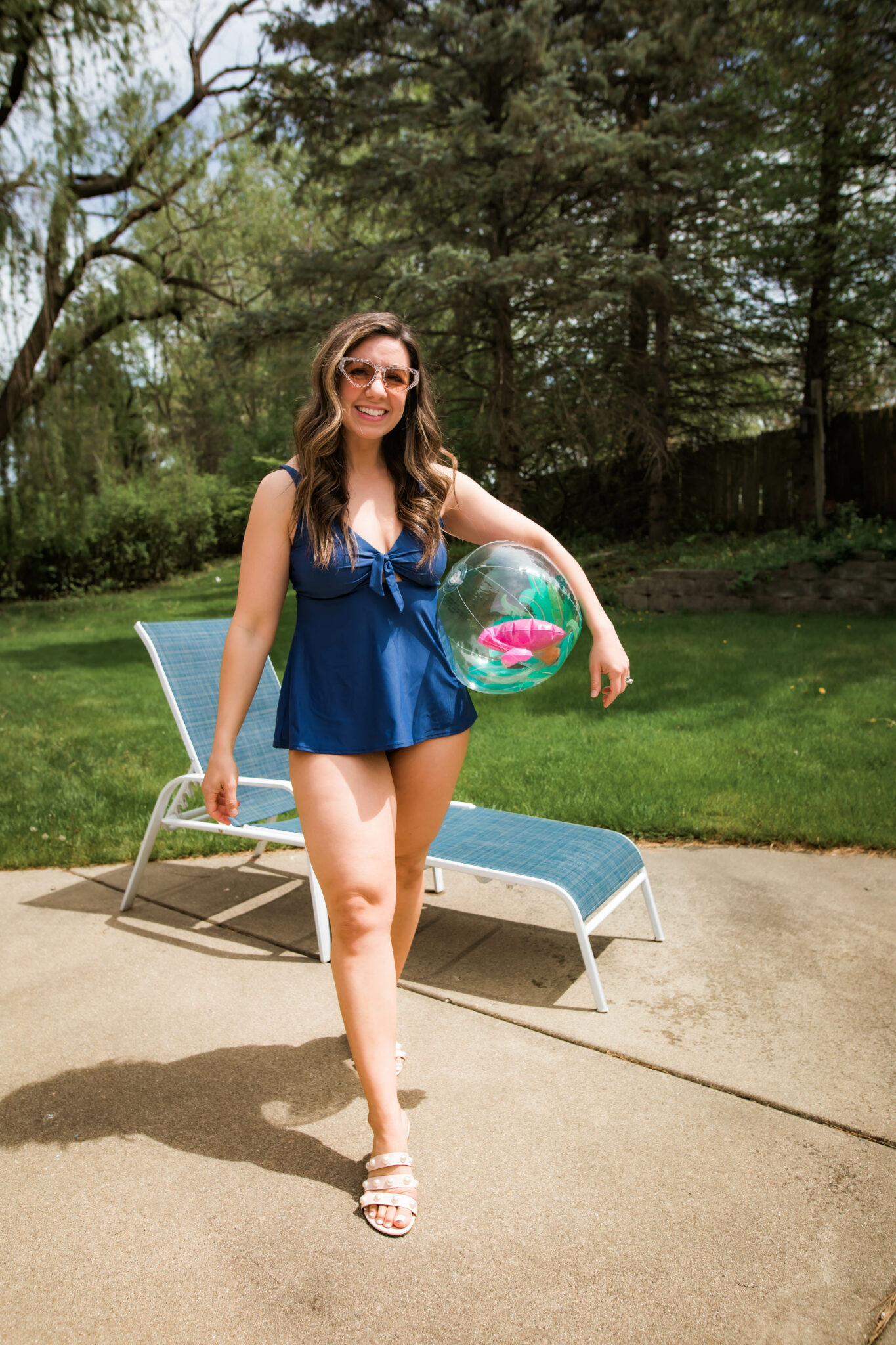 Best Swimsuits for Mom by popular Chicago fashion blog, Glass of Glam: image of a woman holding a beach ball and wearing a blue Bare Necessities two piece with white strap slide sandals. 